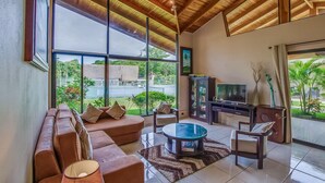 A spacious living room with a large window, a ceiling fan, a sofa set, a coffee table, and a TV. - Hidden Palms Nosara (Nosara)
