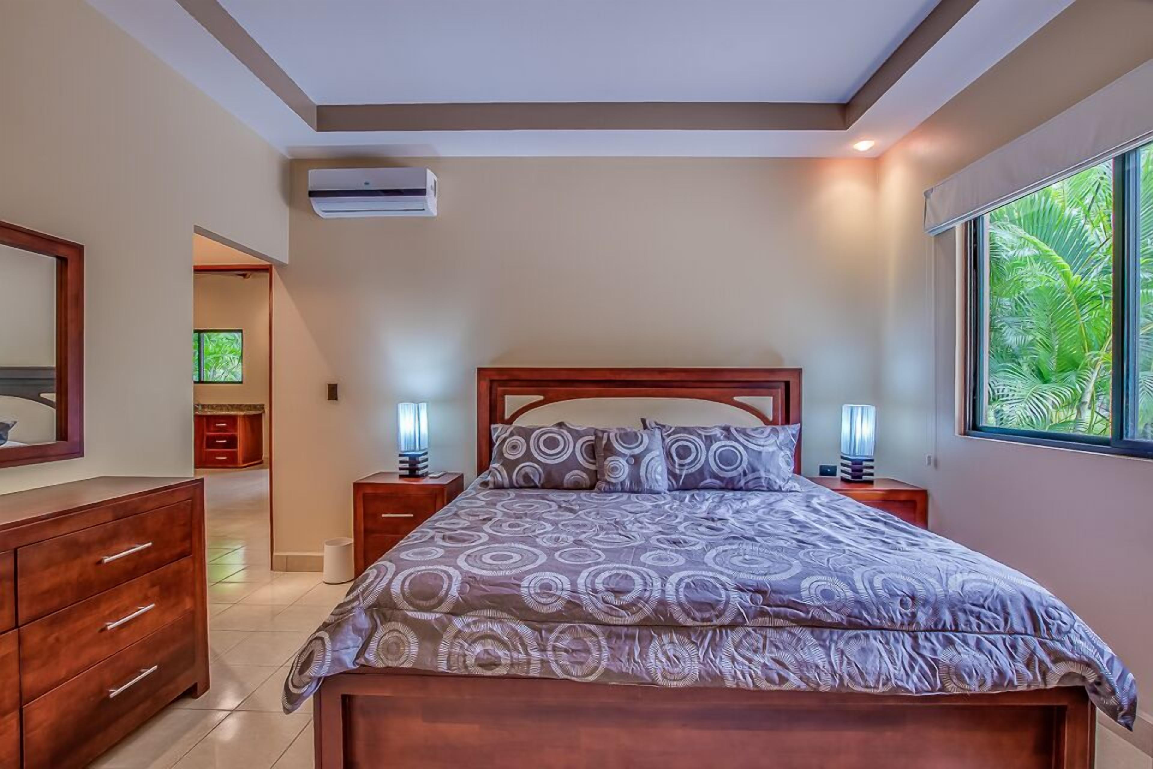 A bedroom with a wooden bed, two bedside tables with lamps, a large window with a view of greenery, and an air conditioning unit on the wall.
