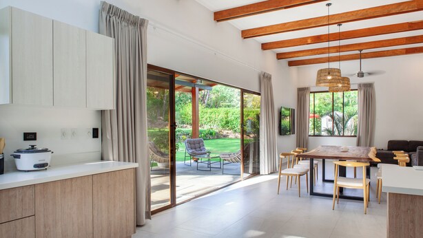 A modern kitchen with a large island, sliding glass doors leading to a patio, and a dining area with a wooden table and chairs. - Hidden Palms Nosara (Nosara)