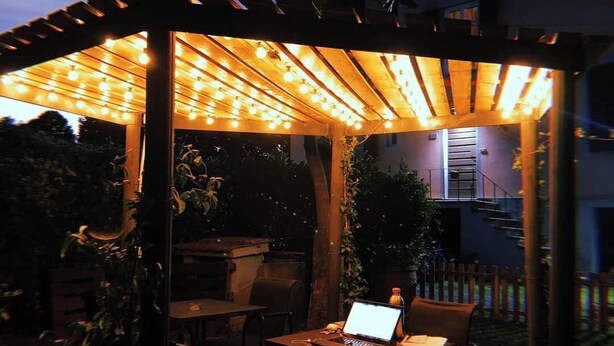 Pátio externo com pérgola coberta por luzes de Natal, um laptop sobre uma mesa e cadeiras.