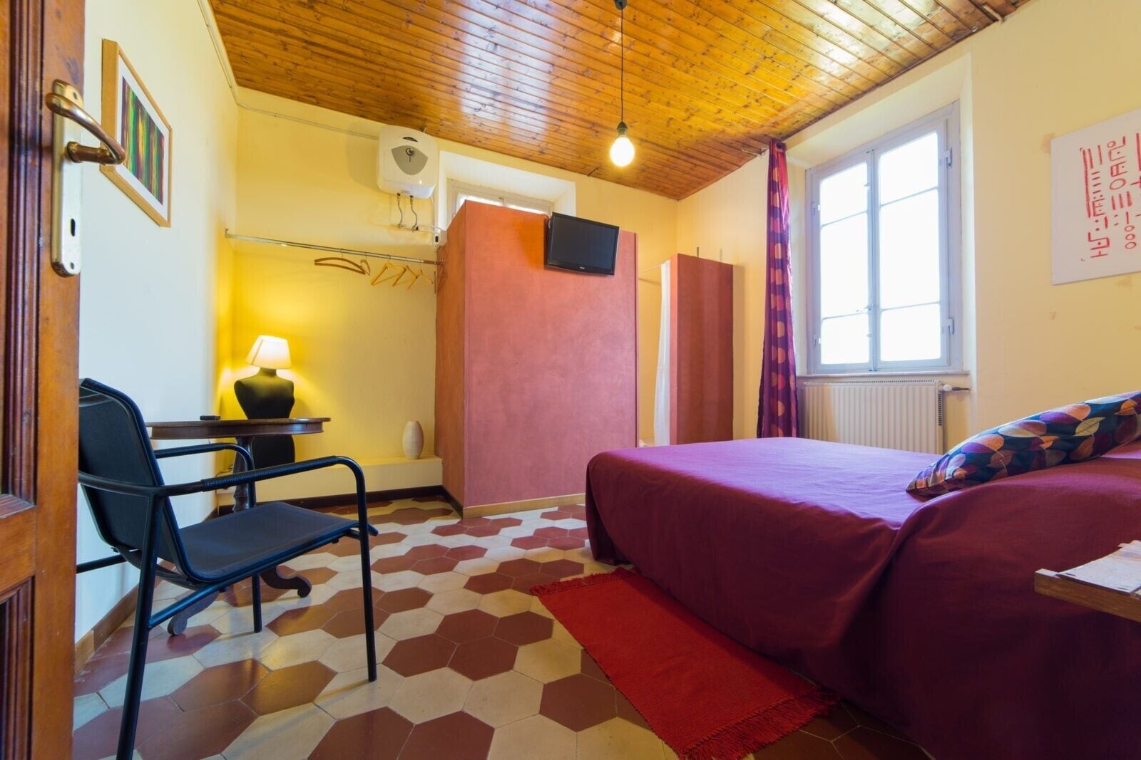 A room with a bed, a desk with a chair, a lamp, a window, and a wooden ceiling.