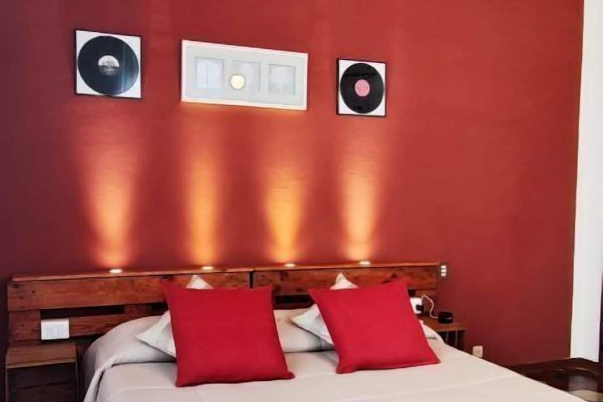 Un dormitorio con techo de madera, pared roja, cama con ropa de cama blanca y roja, y dos altavoces instalados en la pared.