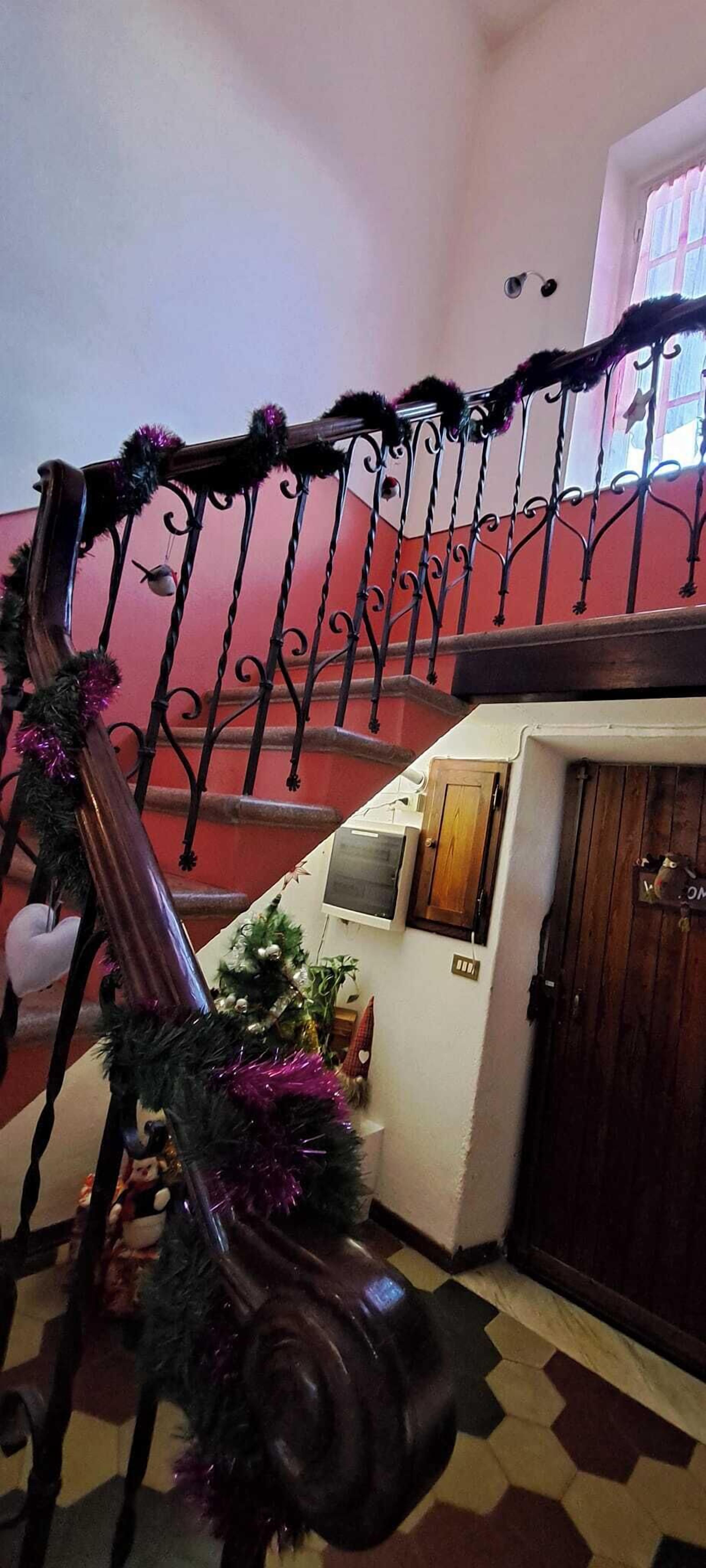 Un escalier orné d’une rampe en fer forgé décorée de guirlandes et de festons, menant à un palier avec une porte en bois et un petit sapin de Noël.