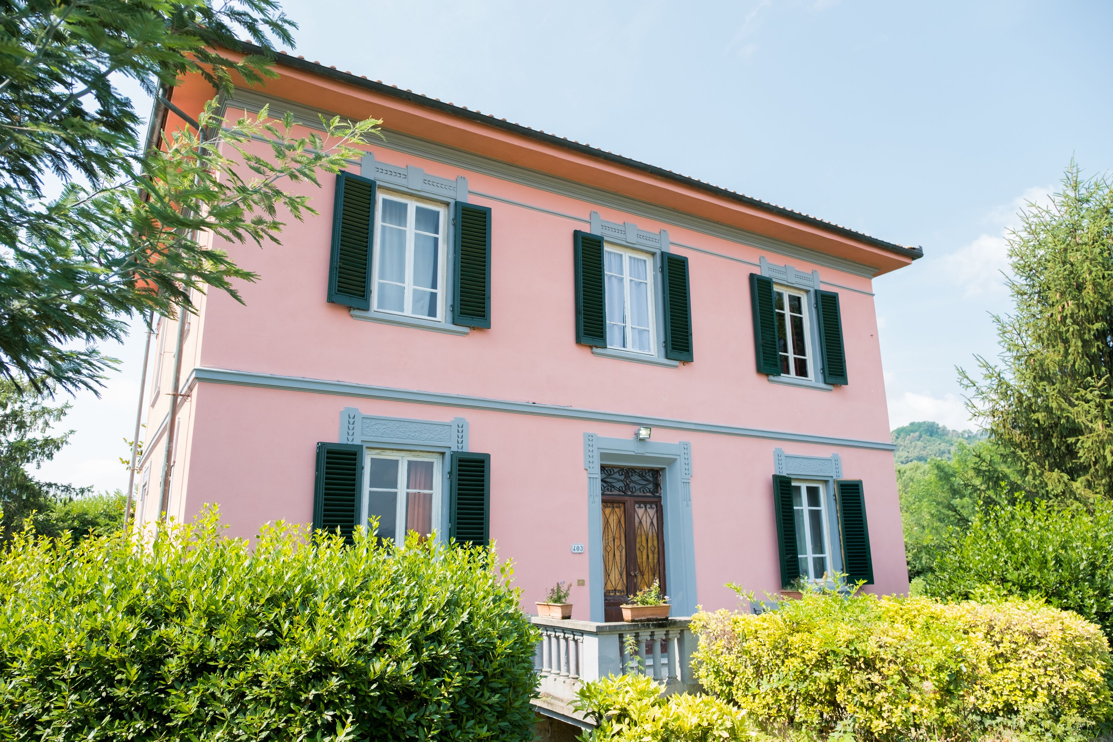 A pink building with green shutters, a balcony, and lush greenery.