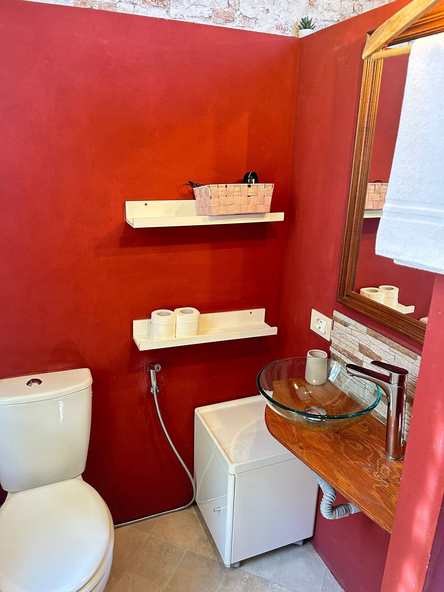 Ein kleines Badezimmer mit roter Wand, weißer Toilette, Waschbecken aus Glas und einem Holzwaschtisch.