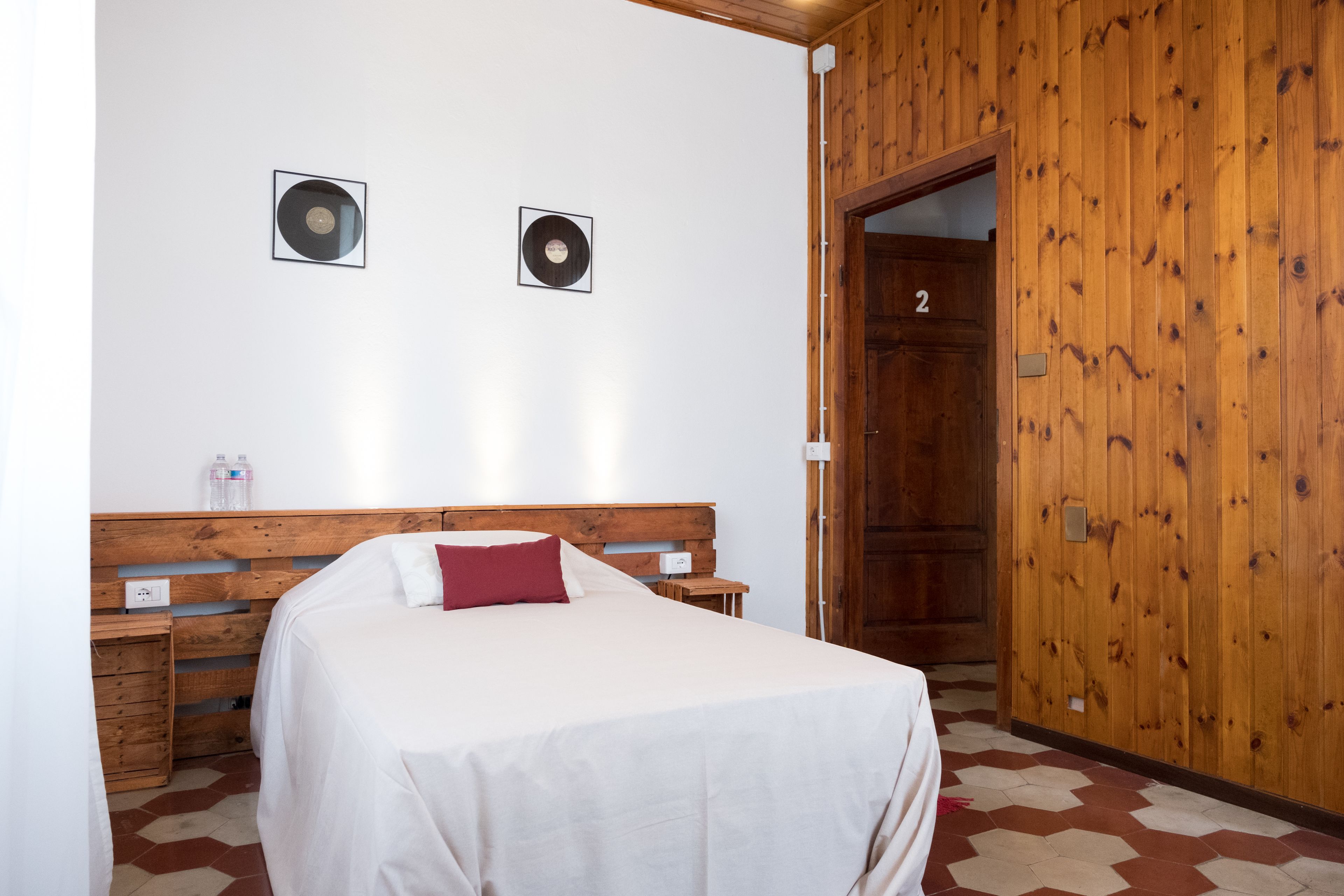 A bedroom with a wooden headboard, two framed vinyl records on the wall, a bed with white bedding and a red pillow, a wooden wardrobe, and a door labeled "2".
