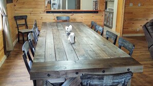 Un comedor rústico de madera con una mesa grande y sillas, una lámpara de candelabro y un refrigerador al fondo.