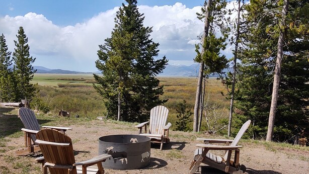 Un espacio al aire libre rústico con sillas Adirondack y una chimenea, rodeado de altos pinos y una vista panorámica de montañas distantes.