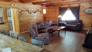Interior acogedor de una cabaña de troncos, con una mesa de comedor de madera, dos sofás y un reloj en la pared.
