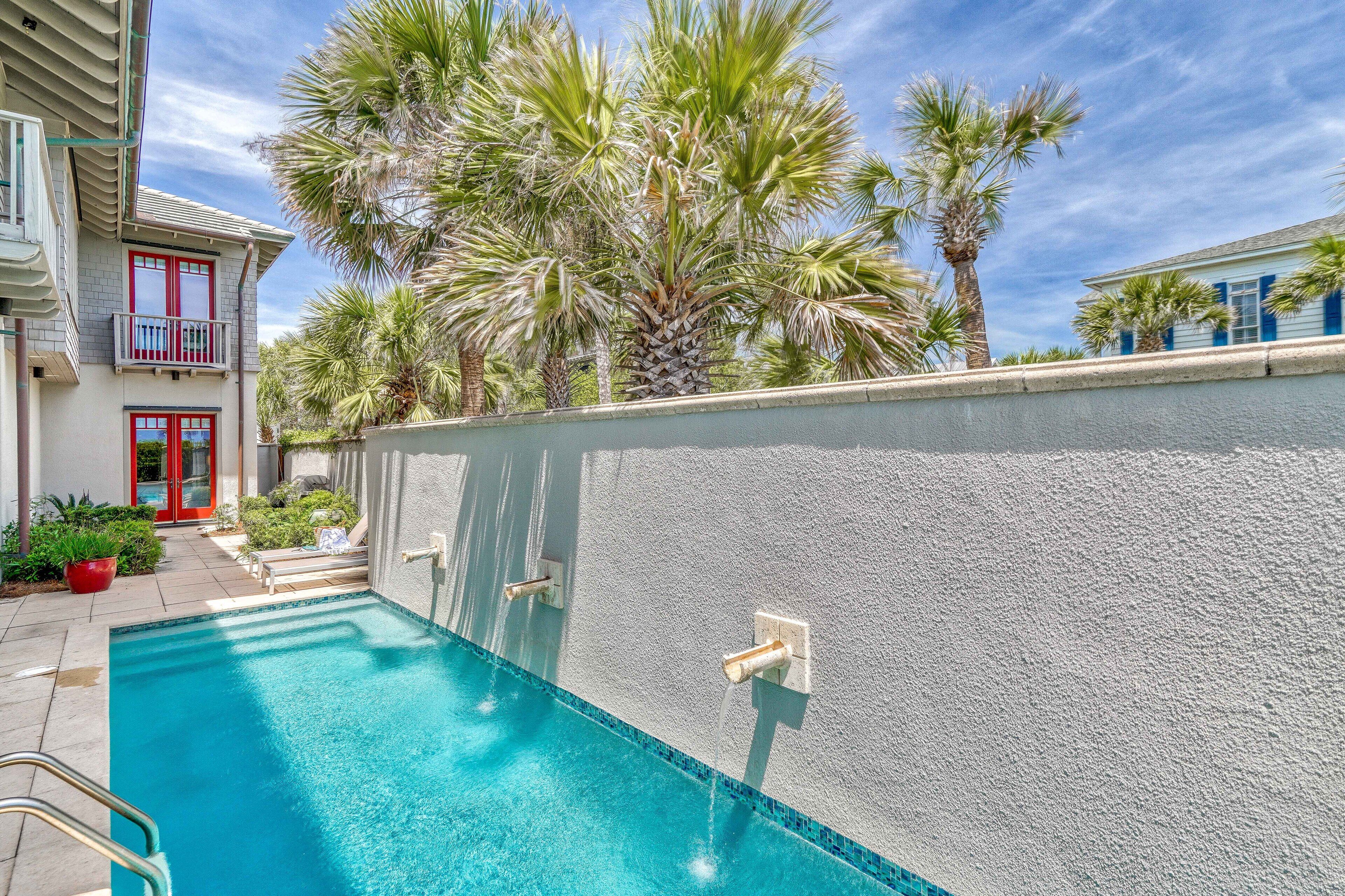 Une piscine avec un mur blanc et une porte rouge, entourée de palmiers et dotée d’un balcon.