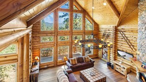 A log cabin interior with a large sofa, a fireplace, and a chandelier. - Grand Cameron Estate by Destination Big Bear : Movie Theater! HOT TUB! Lake Views! PET FRIENDLY 🐾!Walk to the Village! Game Room w/ Pool Table! (Big Bear Lake)