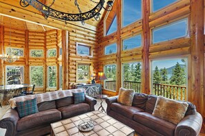 A log cabin interior with a dining area, two sofas, a coffee table, and large windows offering scenic views.