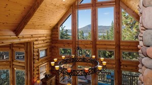 A log cabin interior with a large chandelier, multiple windows offering scenic views, and a wooden ceiling. - Grand Cameron Estate by Destination Big Bear : Movie Theater! HOT TUB! Lake Views! PET FRIENDLY 🐾!Walk to the Village! Game Room w/ Pool Table! (Big Bear Lake)