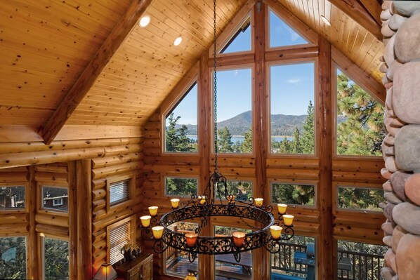 A log cabin interior with a large chandelier, multiple windows offering scenic views, and a wooden ceiling.