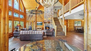 A log cabin interior with a fireplace, a dining area, a staircase, and a chandelier. - Grand Cameron Estate by Destination Big Bear : Movie Theater! HOT TUB! Lake Views! PET FRIENDLY 🐾!Walk to the Village! Game Room w/ Pool Table! (Big Bear Lake)