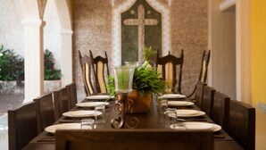 Taula de menjador per a sis amb cadires, un ramader decoratiu amb verdura i una paret amb una creu.