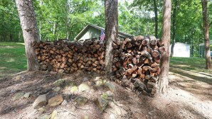 Une pile de bois de chauffage appuyée contre un arbre, un petit abri surmonté d’un drapeau américain et une zone herbeuse avec des arbres en arrière-plan.