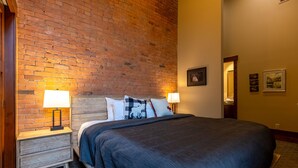 Ein Hotelzimmer mit Backsteinwand, einem hölzernen Kopfteil, einem großen Bett mit dunkelblauer Bettdecke, zwei Nachttischlampen und einem kleinen Nachttisch mit einer Lampe.