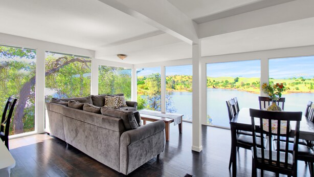 A spacious living area with a large window overlooking a lake, a grey sofa, and a dining table with chairs. - Tulloch Lake House! Panoramic Views & Private Dock (Jamestown)