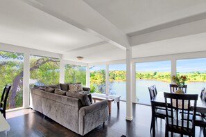 A spacious living area with a large window overlooking a lake, a grey sofa, and a dining table with chairs.