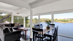 A modern living room with a large window overlooking a lake, a sofa set, and a dining table with chairs. - Tulloch Lake House! Panoramic Views & Private Dock (Jamestown)