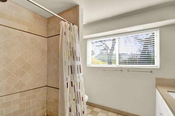 A bathroom with beige tiles, a shower curtain, a window with blinds, and a toilet.