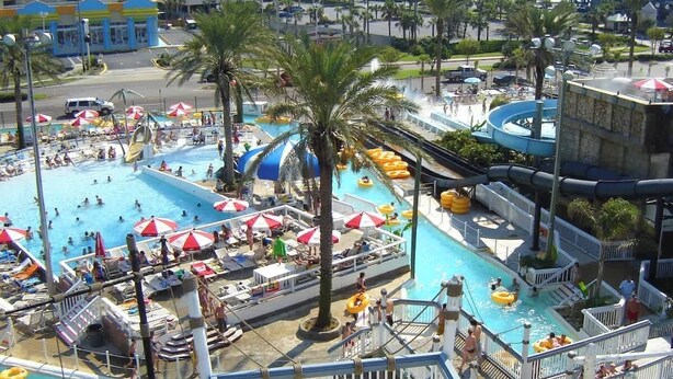 A large outdoor swimming pool with multiple lanes, palm trees, and umbrellas.