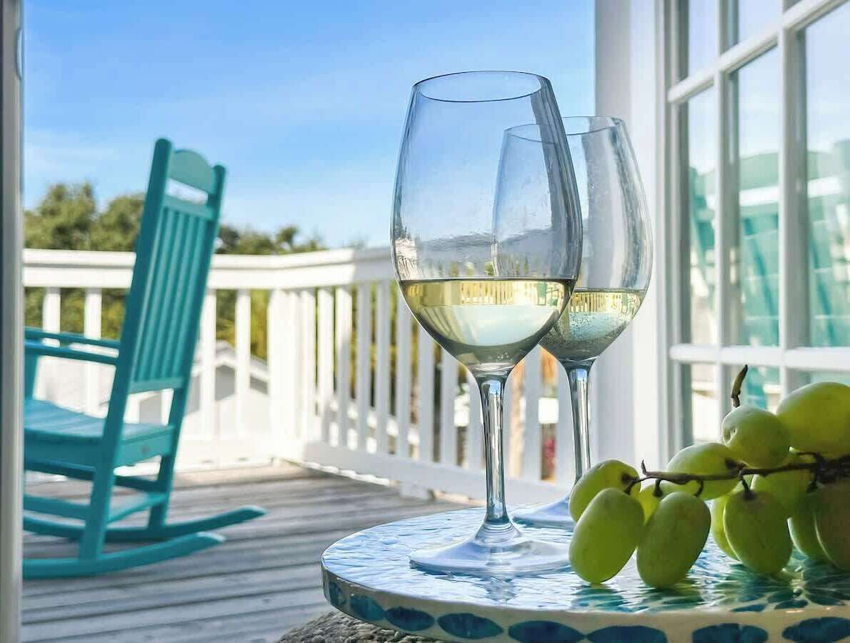 Two wine glasses on a table with a view of a white railing and a blue chair.