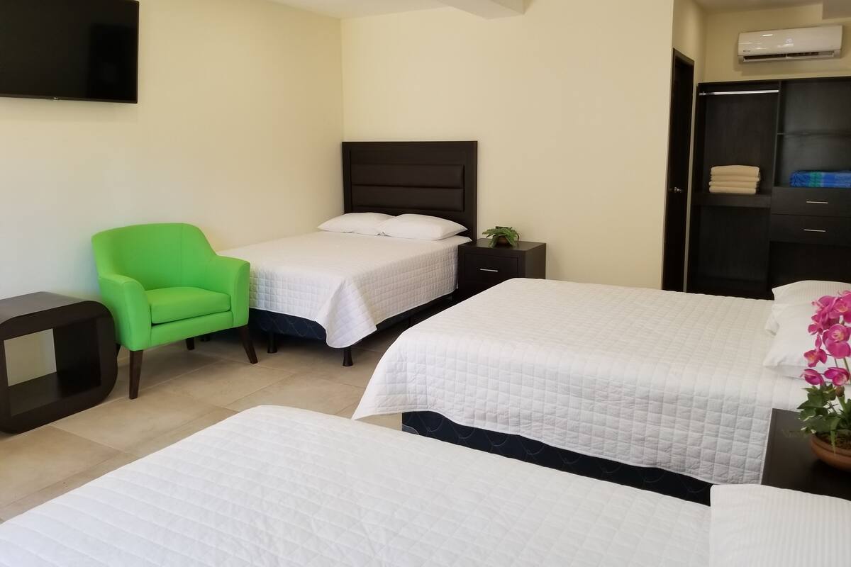 A hotel room with two beds, a green armchair, a TV, a bedside table, and an air conditioning unit.
