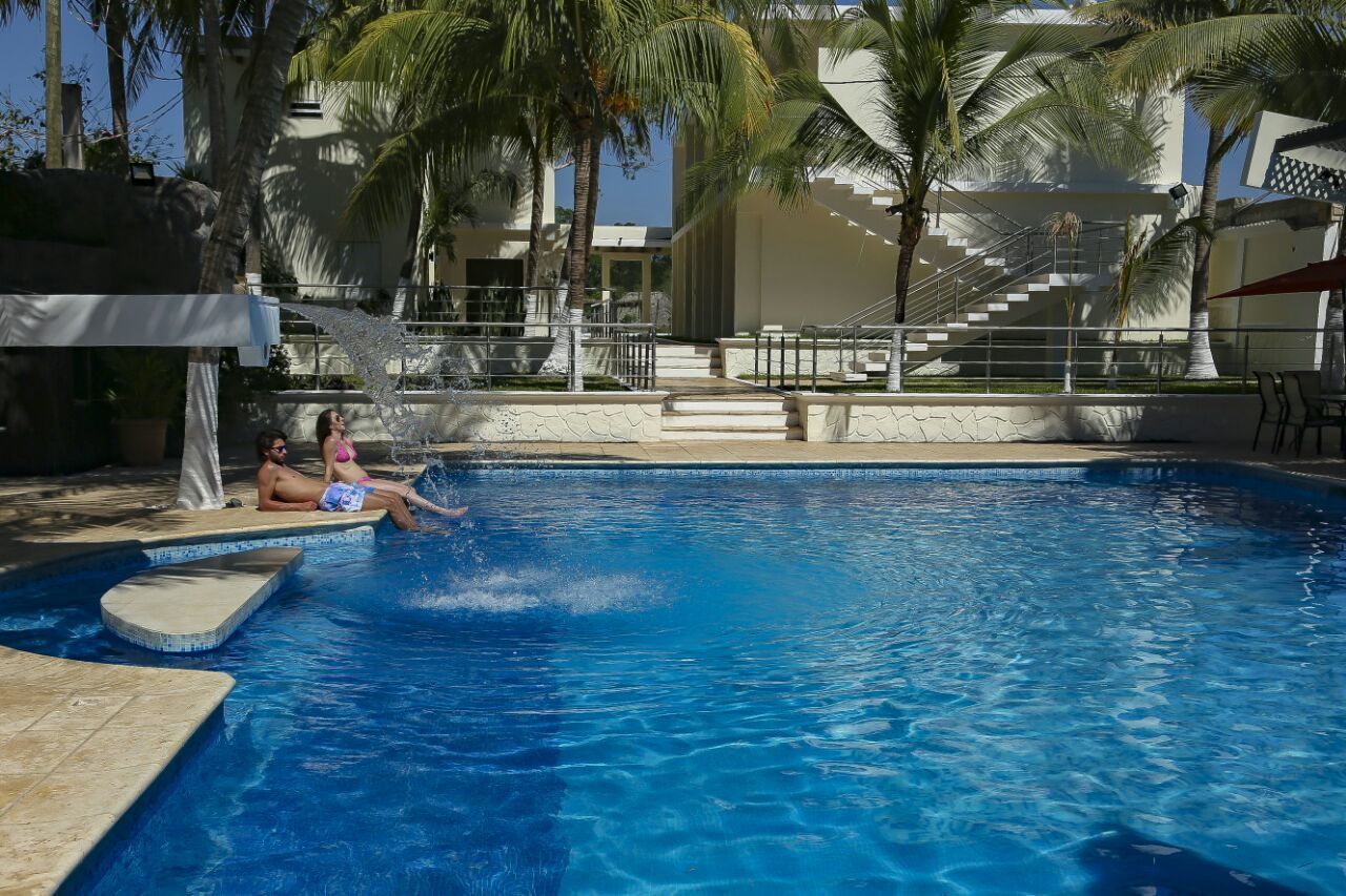A swimming pool with two people relaxing on the edge, surrounded by palm trees and a modern building with a staircase.
