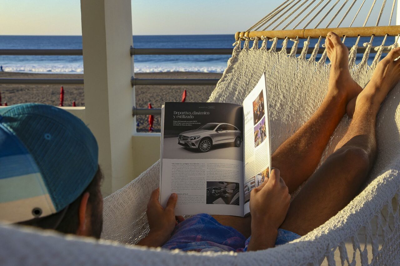 Una persona recostada en una hamaca leyendo una revista, con una playa y el océano al fondo.