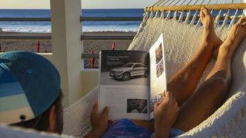 Una persona recostada en una hamaca leyendo una revista, con una playa y el océano al fondo.