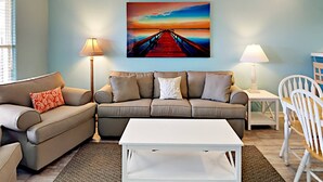 Ein Wohnzimmer mit einem beigen Sofa, einem weißen Couchtisch und einem Gemälde, das einen Sonnenuntergang über einem Steg zeigt.