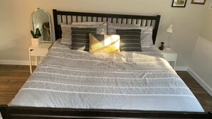 Ein ordentlich bezogenes Bett mit gemustertem Überwurf und Kissen, ein Nachttisch mit Lampe, Spiegel und einer kleinen Topfpflanze.