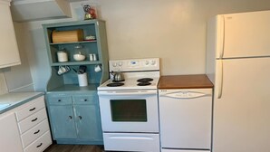 Eine kompakte Küche mit weißem Herd, Backofen und Kühlschrank, einem türkisblauen Schrank mit Küchenutensilien und einer Holz Arbeitsplatte.