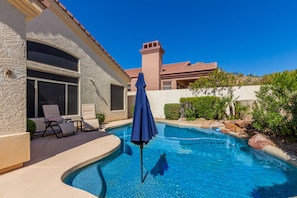 A swimming pool with lounge chairs and an umbrella, surrounded by a landscaped garden and a building with a tiled roof. - Beautiful home w/ private heated pool in quiet gated community. (Mesa)