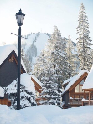 Uma cabana coberta de neve com chalés de madeira, um poste de luz e montanhas ao fundo.