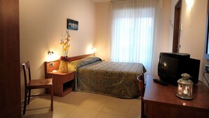 A hotel room with a bed, a desk with a television, a chair, and a nightstand with a lamp and flowers. - Hotel Arma (Taggia)