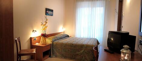 Habitación de hotel con cama, escritorio con televisor, silla y mesita de noche con lámpara y flores.
