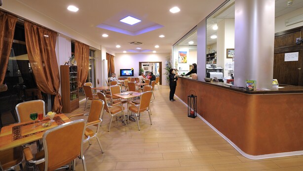 A restaurant with multiple tables and chairs, a counter with a staff member, and a television in the background. - Hotel Arma (Taggia)