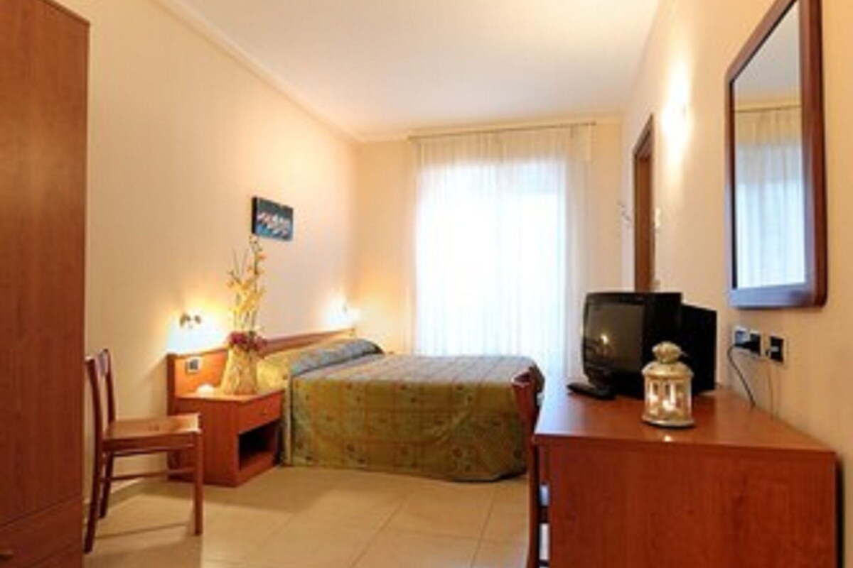 A small, well-lit bedroom with a bed, a desk, a chair, a television, and a mirror.
