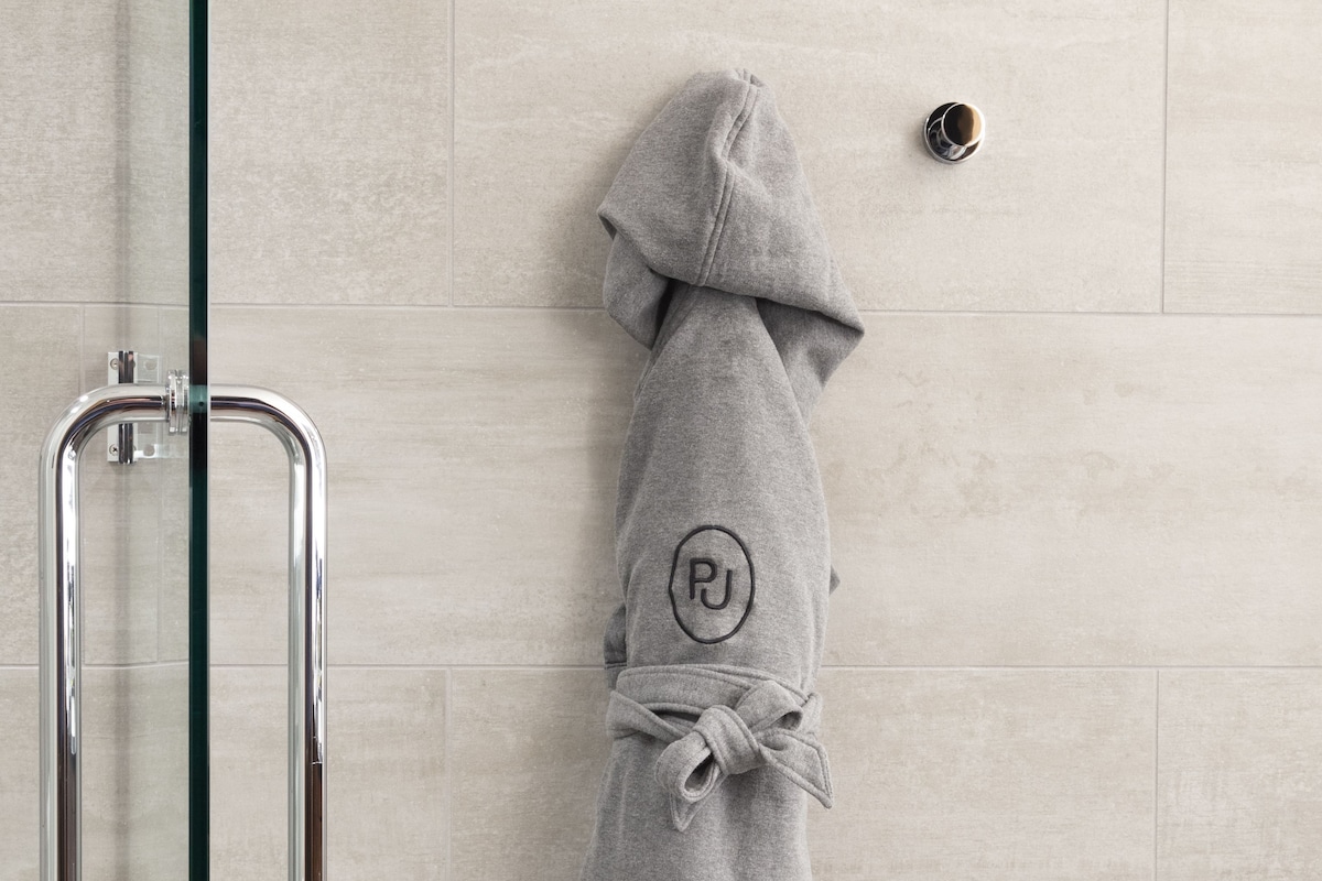Una bata gris con el logo de PJ colgada en una pared de azulejos, una puerta de ducha de cristal y un pasamanos de metal.