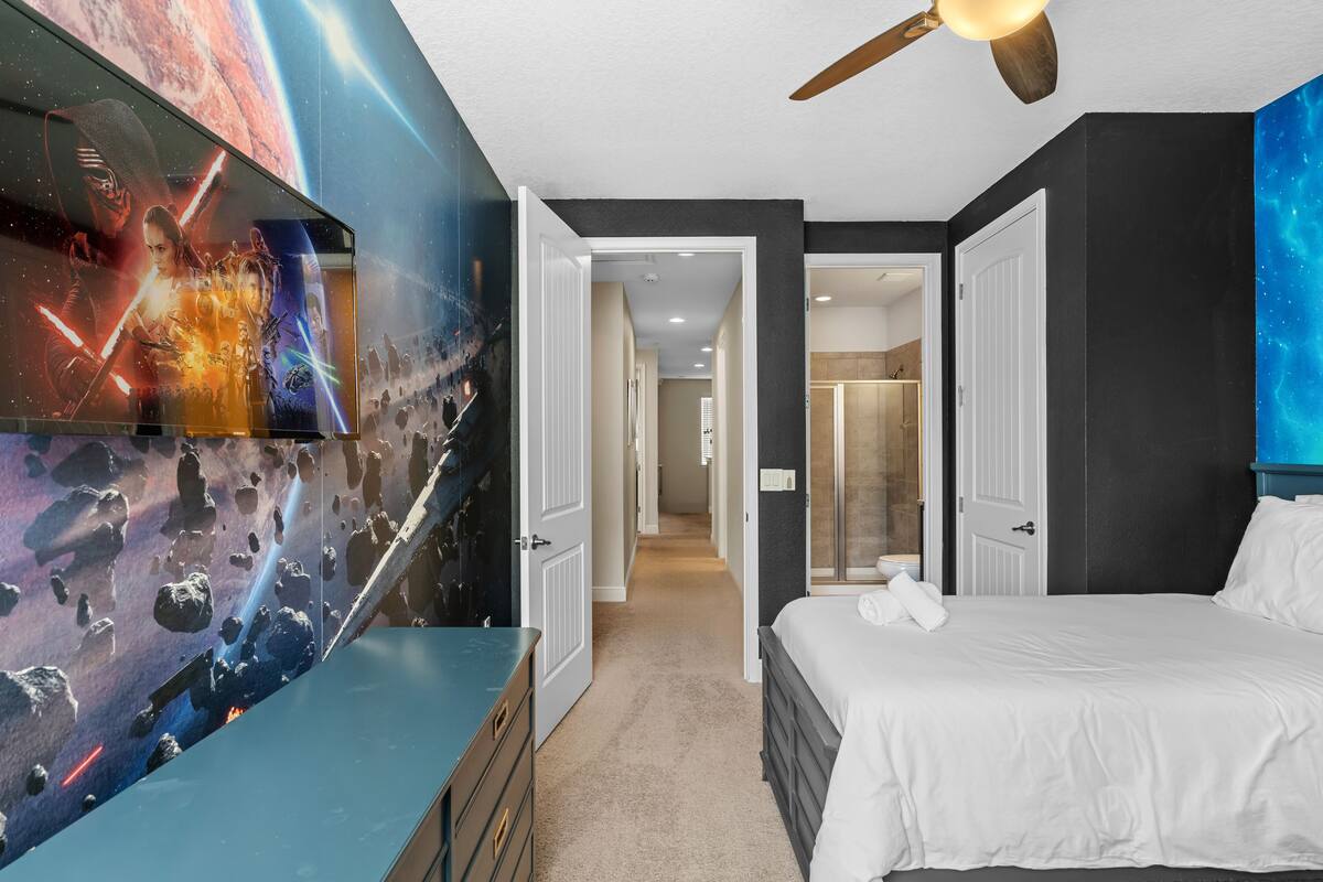 Un dormitorio con una cama, un aparador, un ventilador de techo y una pared con un mural alusivo al espacio.