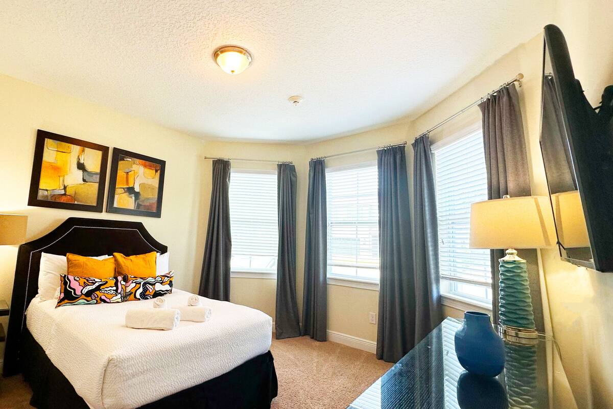 Habitación de hotel con cama, dos lámparas de noche, mesita auxiliar, ventana con cortinas y dos cuadros abstractos en la pared.