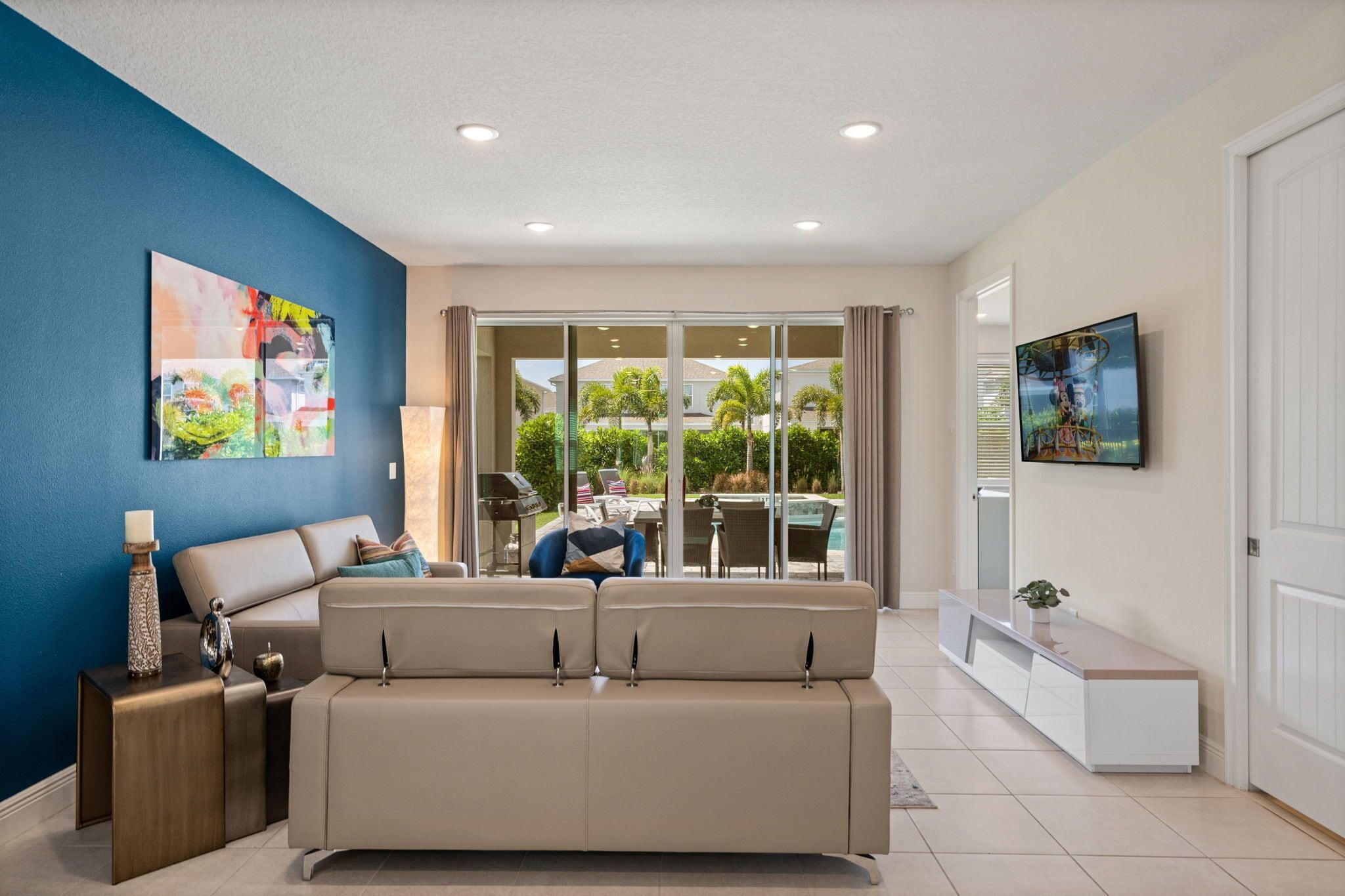 A living room with a beige sofa, a TV mounted on the wall, and a large window with a view of a pool and greenery.
