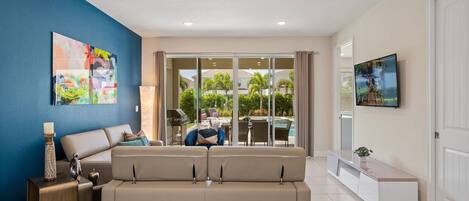 A living room with a beige sofa, a TV mounted on the wall, and a large window with a view of a pool and greenery.