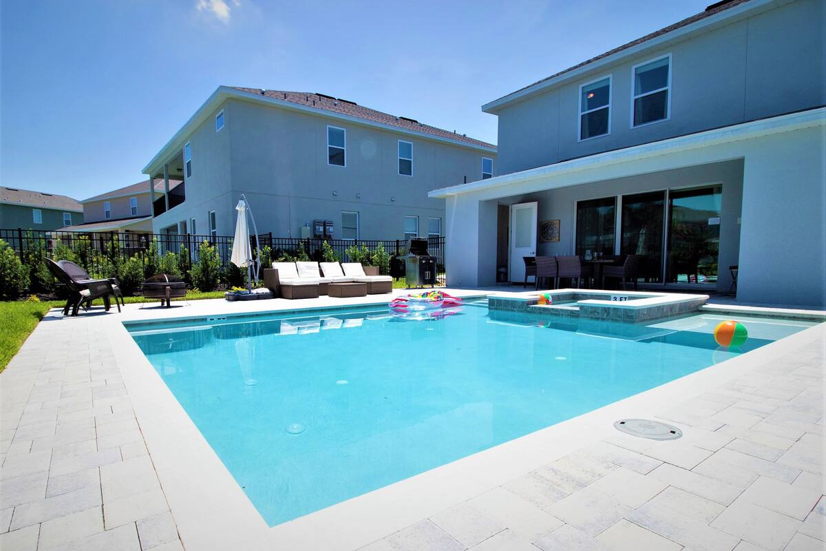 Una piscina rectangular con un borde de baldosas, rodeada de sillas de descanso y una zona de juegos pequeña con juguetes.