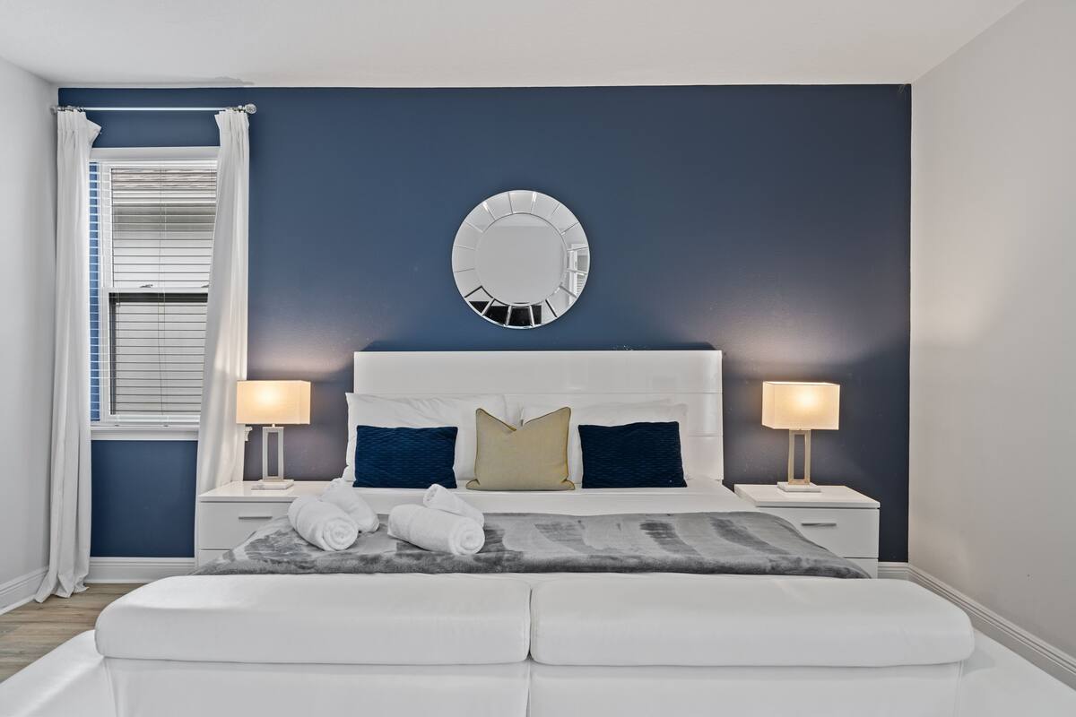 Un dormitorio con una cama blanca, dos mesitas de noche, un espejo redondo y una ventana con cortinas blancas.