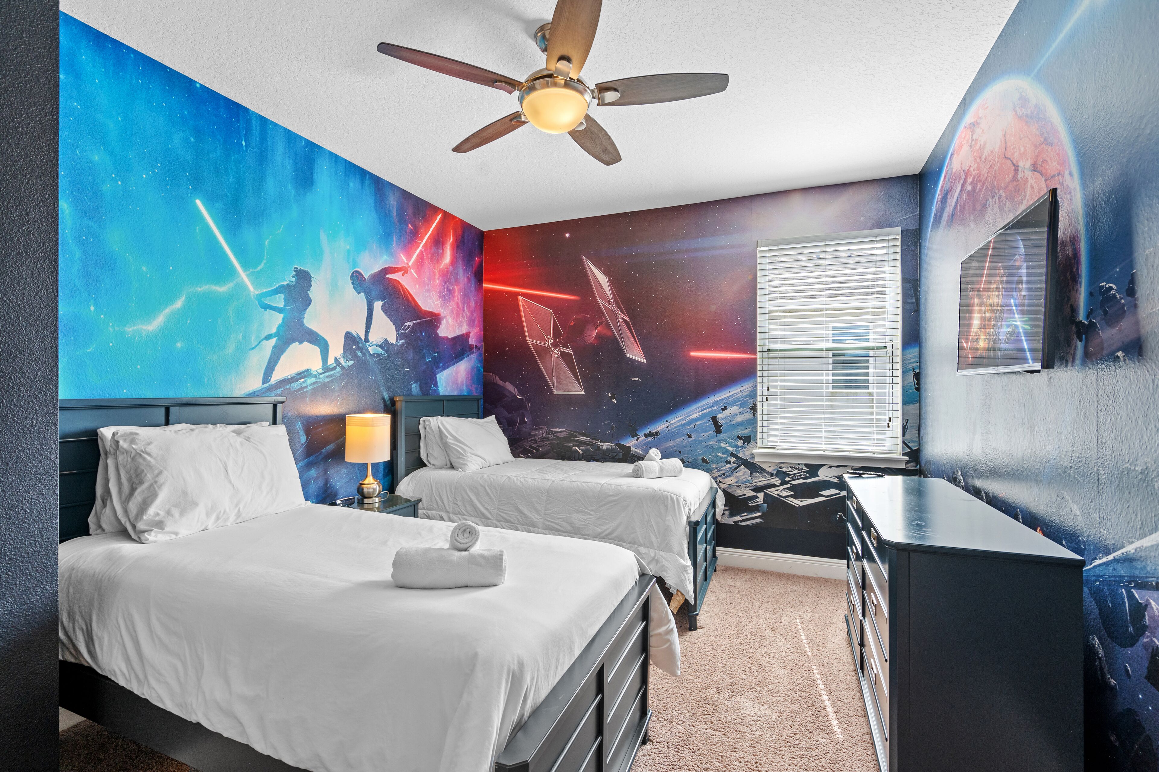 A hotel room with two beds, a ceiling fan, a large wall mural, and a window with blinds.