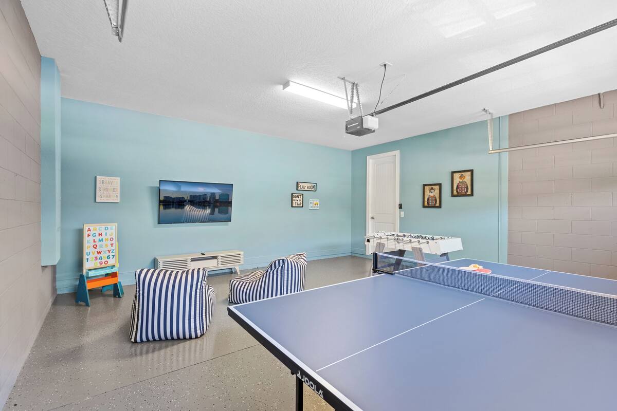 Una habitación con mesa de ping pong, mesa de fútbolín, una pequeña zona de juegos con letras del alfabeto y un cuadro de un paisaje urbano en la pared.